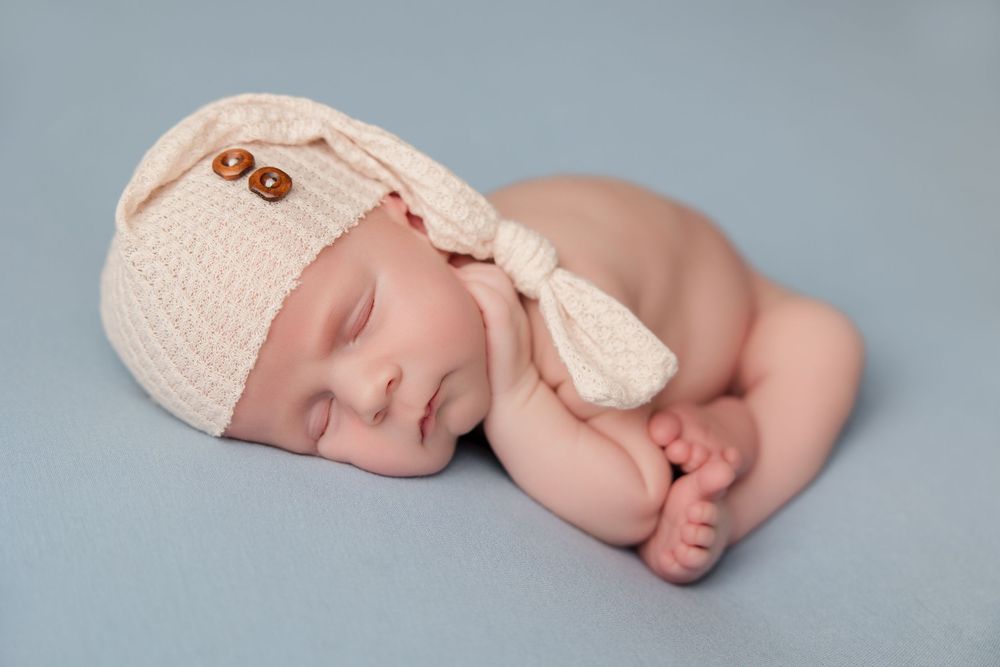 A peaceful sleeping newborn wearing a white knit hat curls up in a soft pose during a professional photography session.