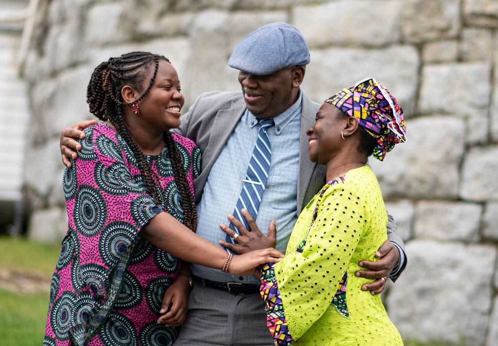 Three joyful people in colorful African attire embracing and laughing outdoors together. Photo by Xenophon Photography in Maine.
