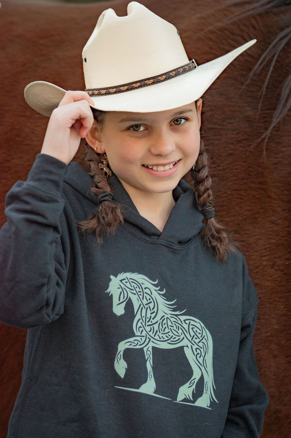 Smiling young girl in white cowboy hat and horse-print hoodie posing near horse. Photo by Xenophon Photography in Maine.