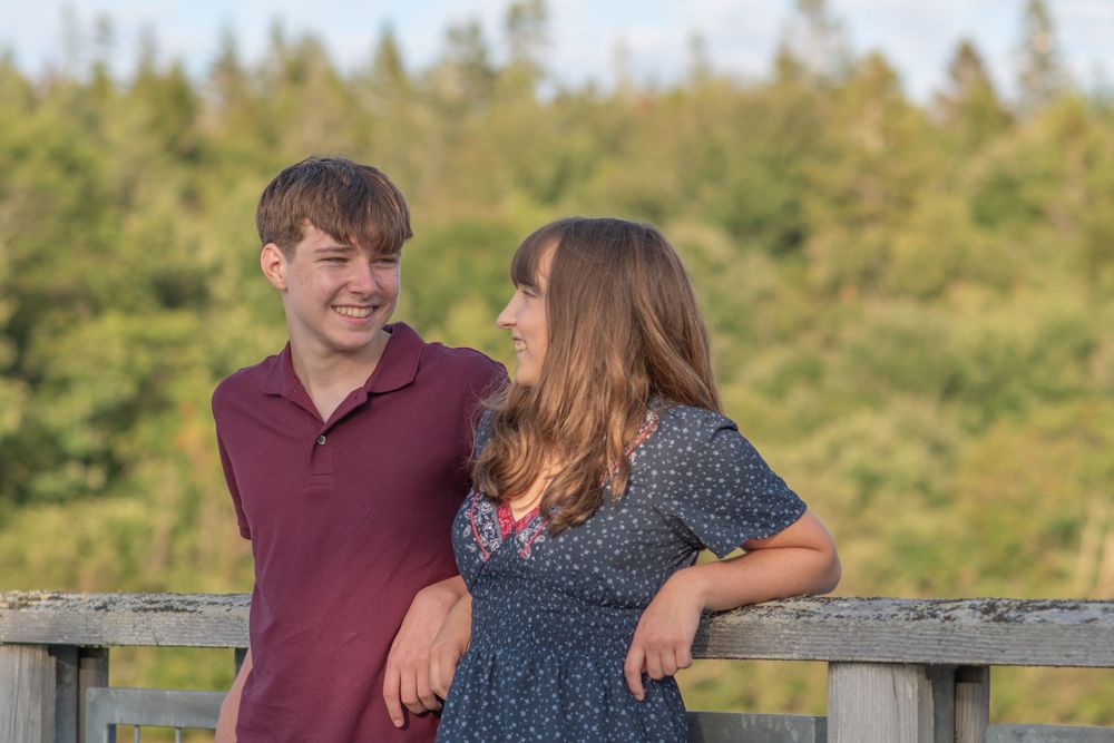 Two teens smile and laugh together on a wooden bridge surrounded by lush green trees outdoors.Photo by Xenophon Photography in Maine.