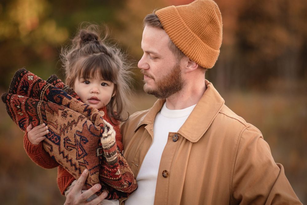 Bearded father in tan coat holding his young daughter in autumn outdoor setting. Photo by Xenophon Photography in Maine.