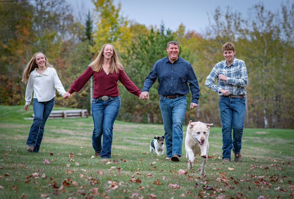 Happy family of four walking dogs through colorful autumn park, smiling and holding hands outdoors. Photo by Xenophon Photography in Maine.
