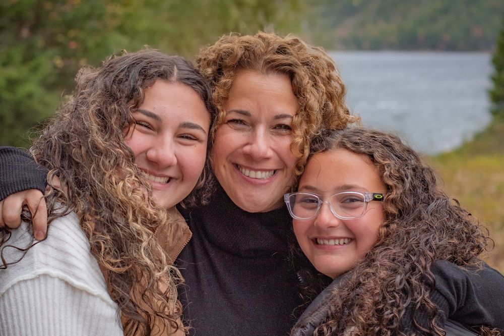 Three curly-haired females, a mother and two daughters, smiling together near a lake. Photo by Xenophon Photography in Maine.
