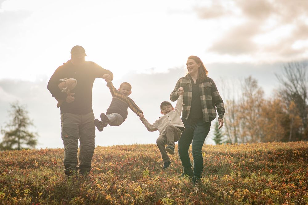Joyful family swinging two children by hands in sunny autumn meadow with colorful trees. Photo by Xenophon Photography in Maine.