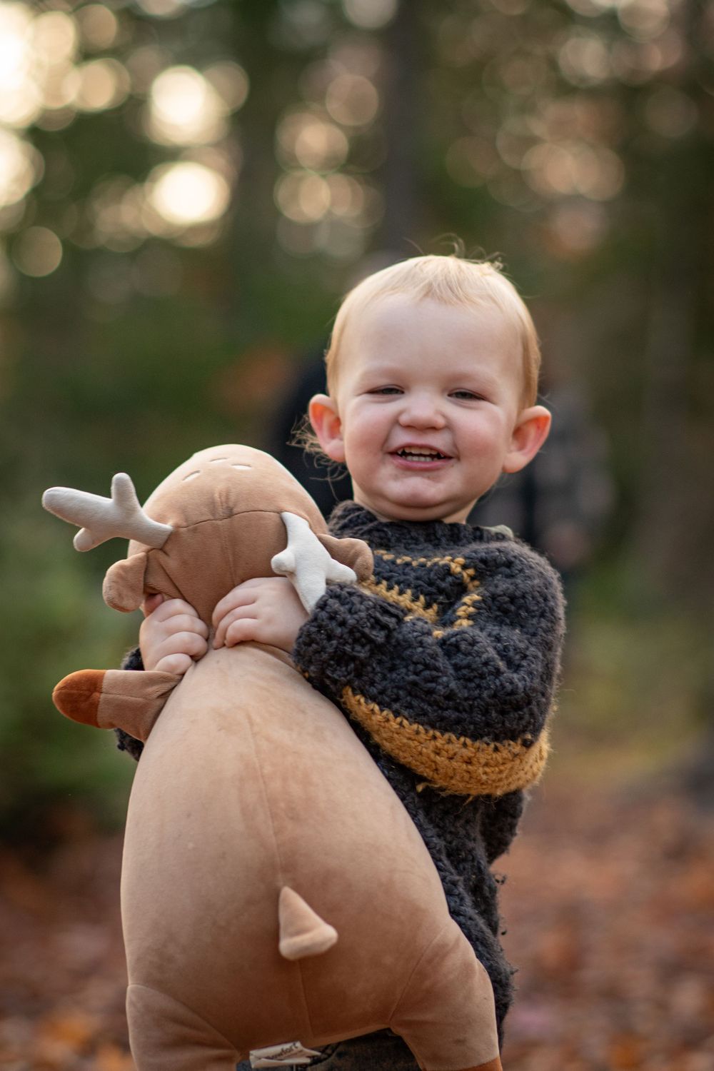 Happy blonde toddler boy hugging large stuffed reindeer toy in autumn forest setting. Photo by Xenophon Photography in Maine.