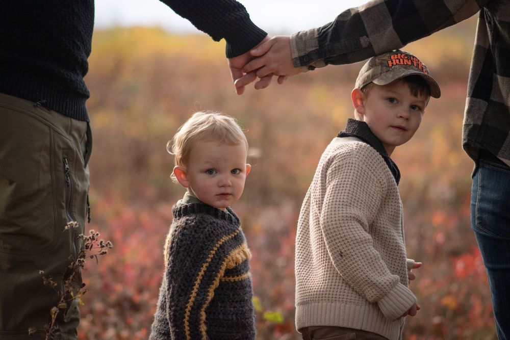 Two young children hold hands with adults in an autumn field with colorful fall foliage. Photo by Xenophon Photography in Maine.