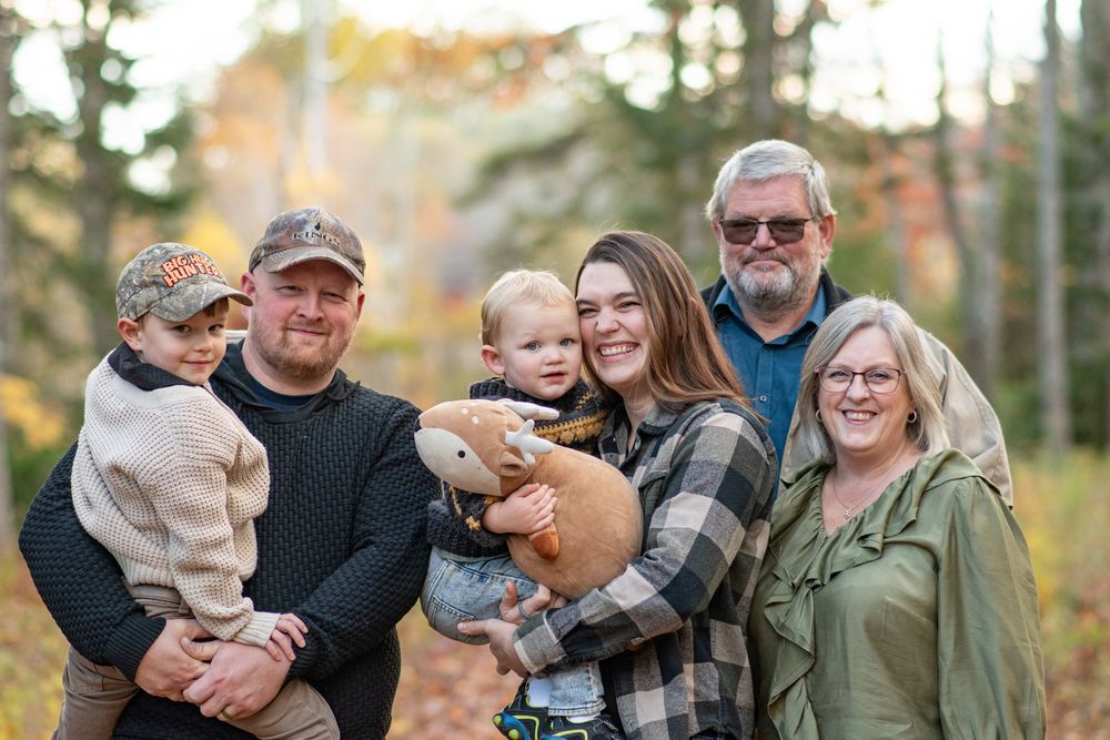 Multi-generational family of six smiling outdoors among colorful fall foliage with baby and stuffed animal. Photo by Xenophon Photography.