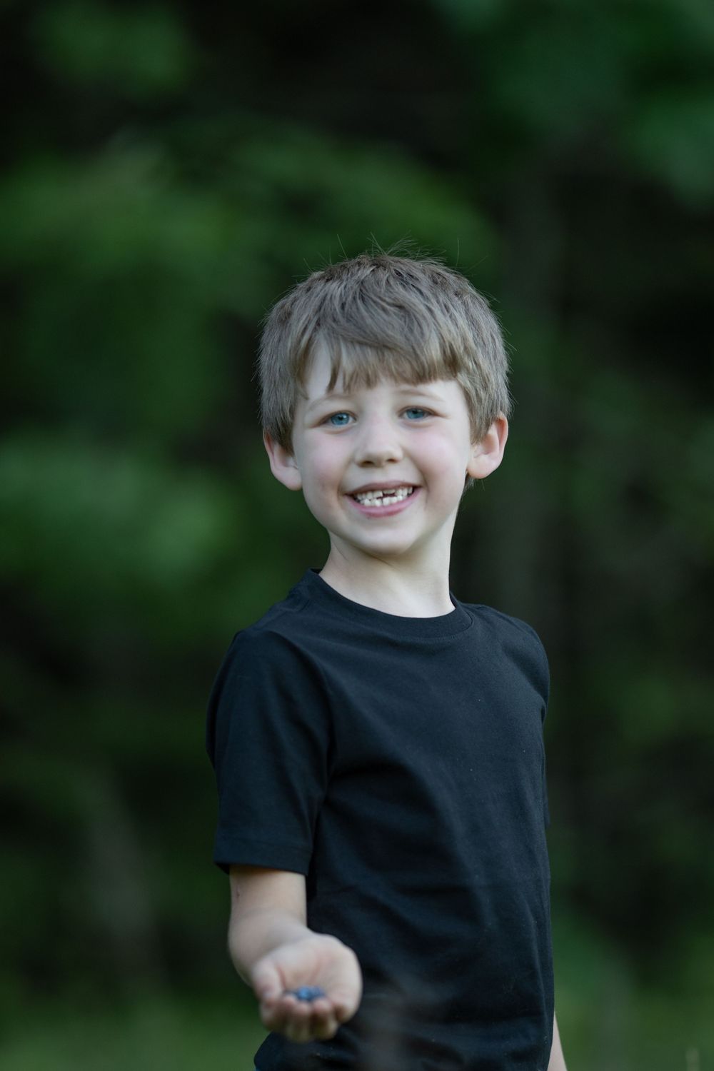 Smiling young boy in black t-shirt reaching out his hand outdoors with green trees in background. Photo by Xenophon Photography in Maine.