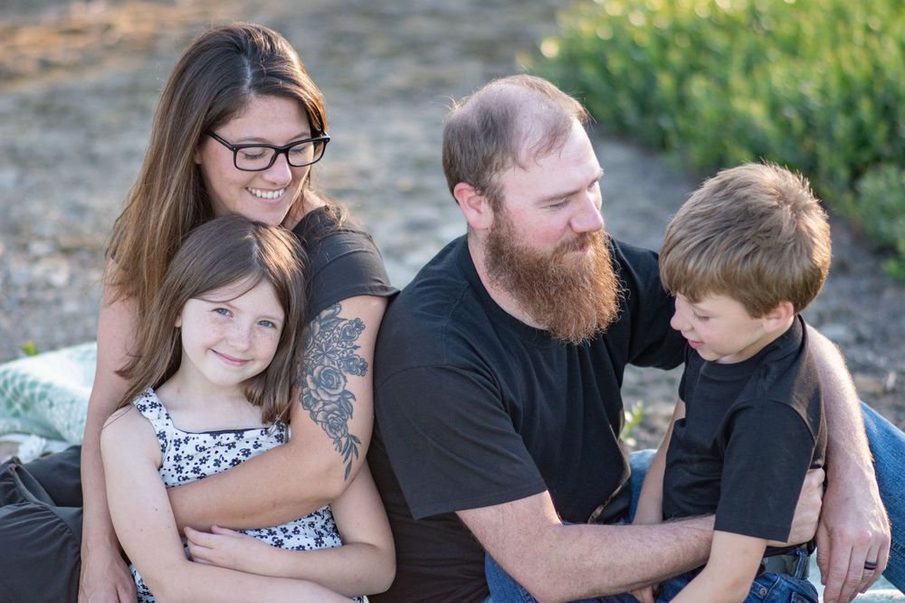 Family of four with tattooed parents and two young children sitting together outdoors in soft light. Photo by Xenophon Photography in Maine.