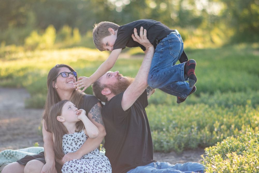 Bearded father lifting laughing son in air while mother and daughter watch outdoors in golden light. Photo by Xenophon Photography in Maine.