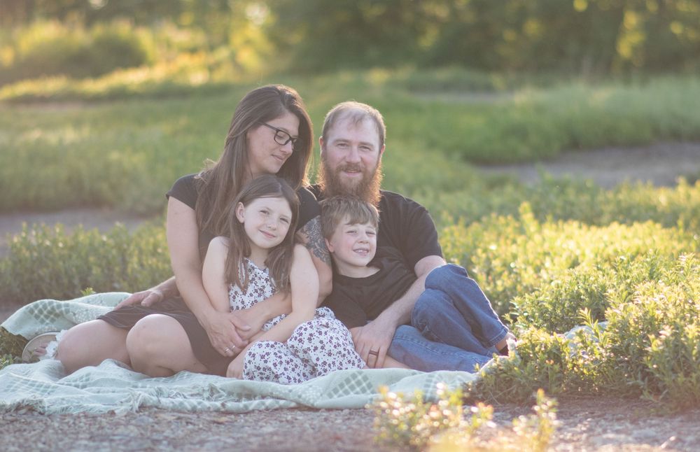 Family of four sitting together on blanket in sunny field, smiling warmly in golden hour light. Photo by Xenophon Photography in Maine.