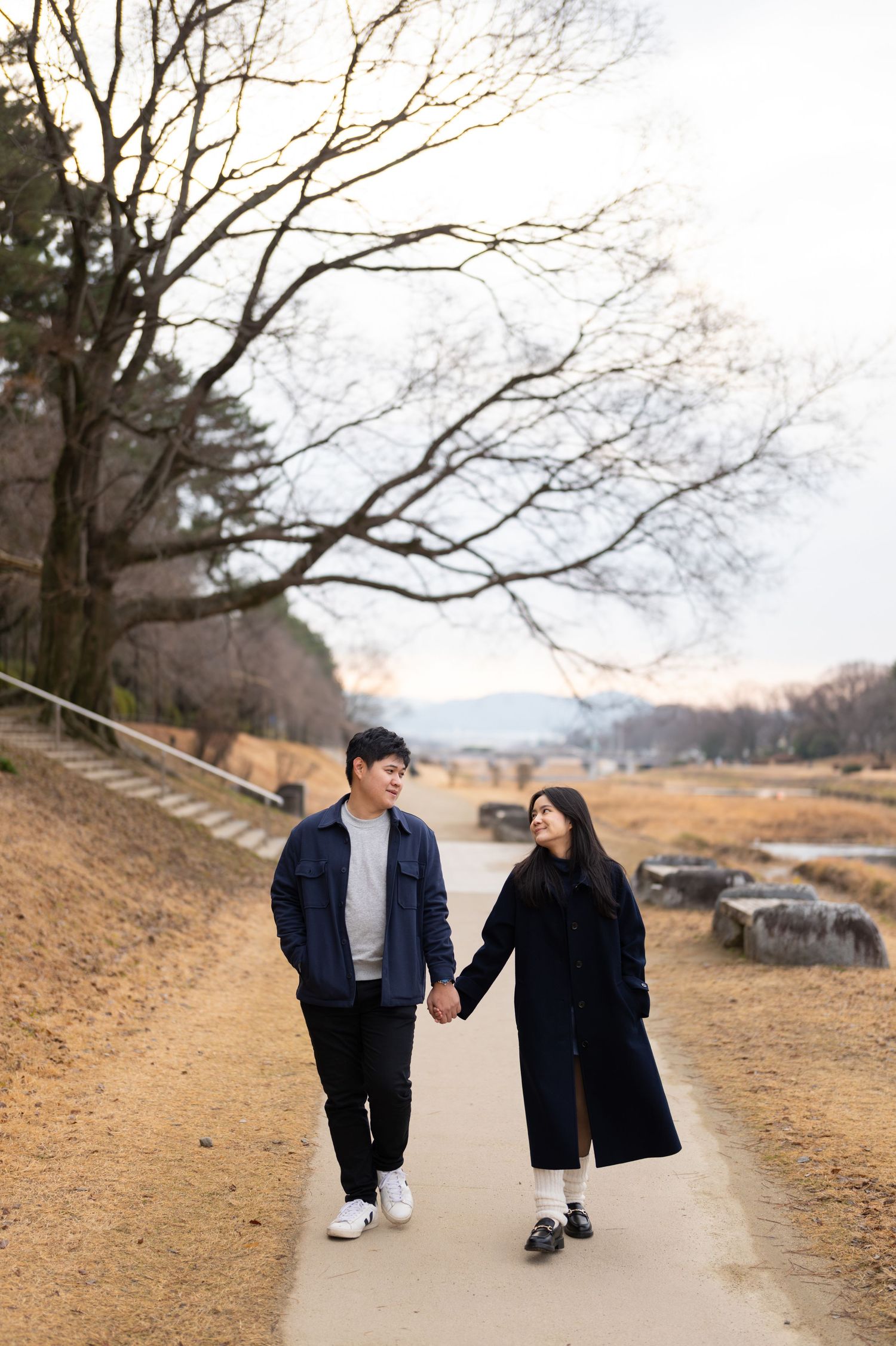 Young couple holding hands and walking along a scenic park path lined with bare winter trees.
