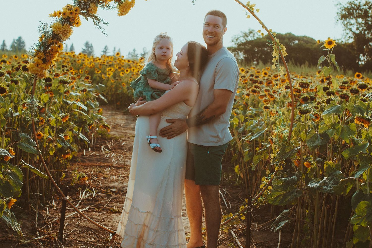 A couple stands together in a golden sunflower field during sunset while holding a mason jar and drinks.