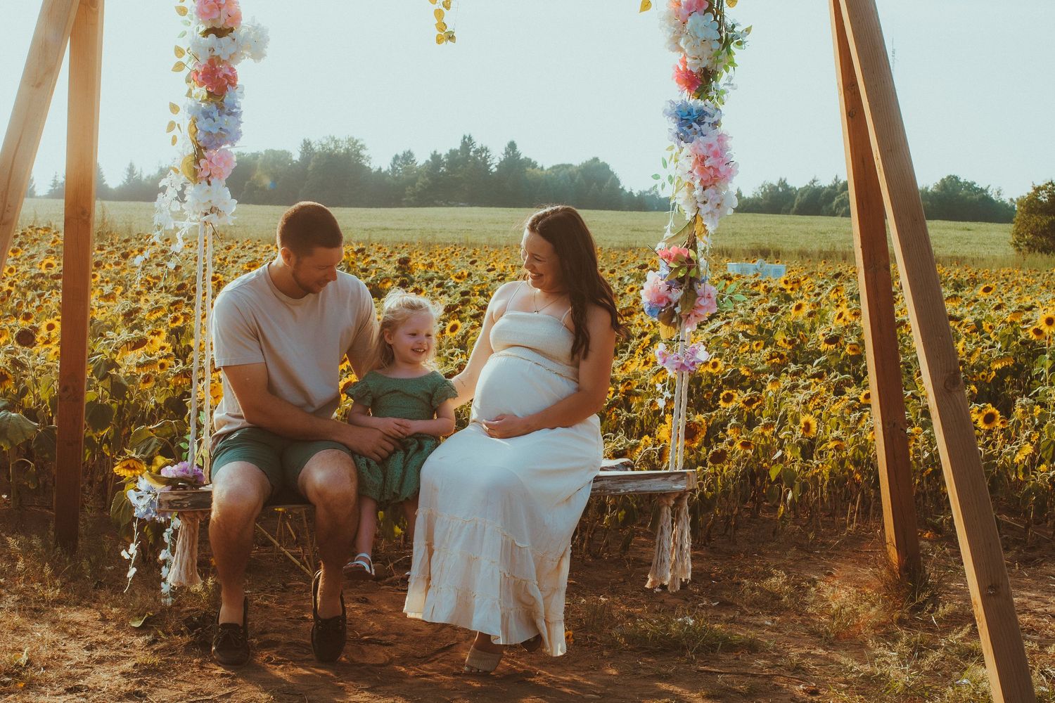 A family poses together at sunset in a sunflower field with warm golden lighting and photography equipment visible.