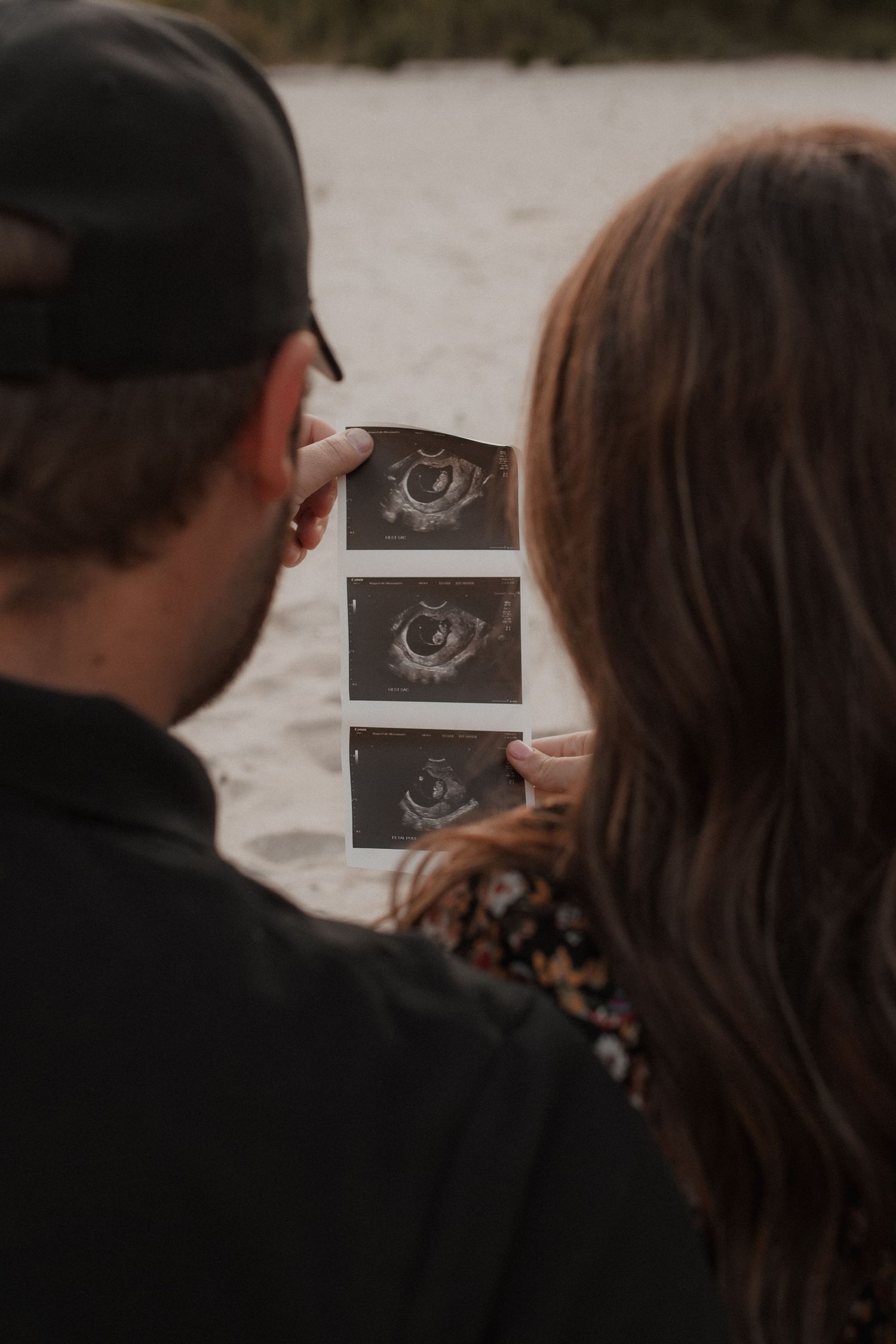 Back view of couple looking at ultrasound pictures together outdoors during pregnancy announcement photoshoot.