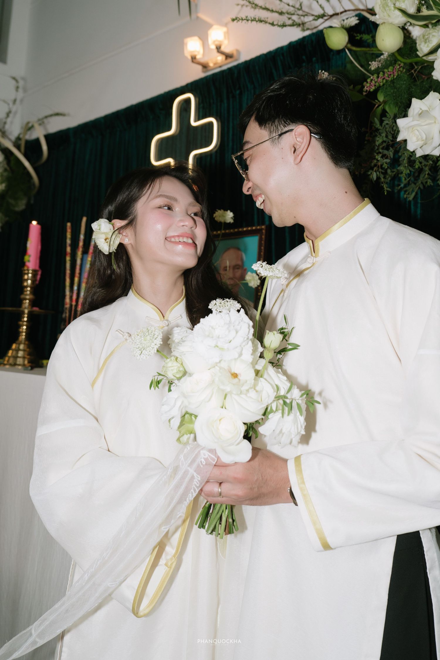 Smiling Vietnamese bride and groom in white ao dai hold white peony bouquet before cross altar.