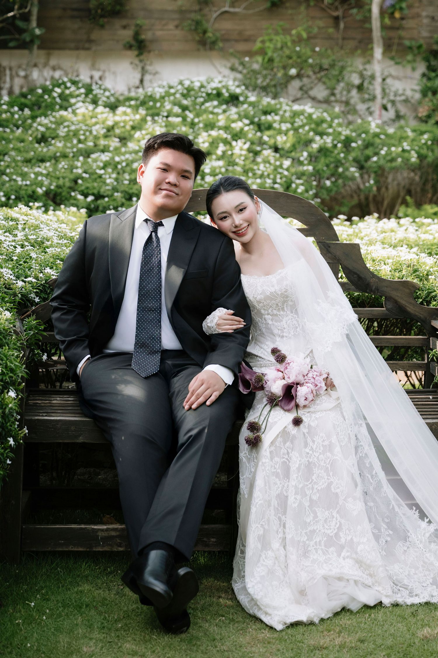 A bride and groom in wedding attire pose on a wooden bench in a lush garden setting.