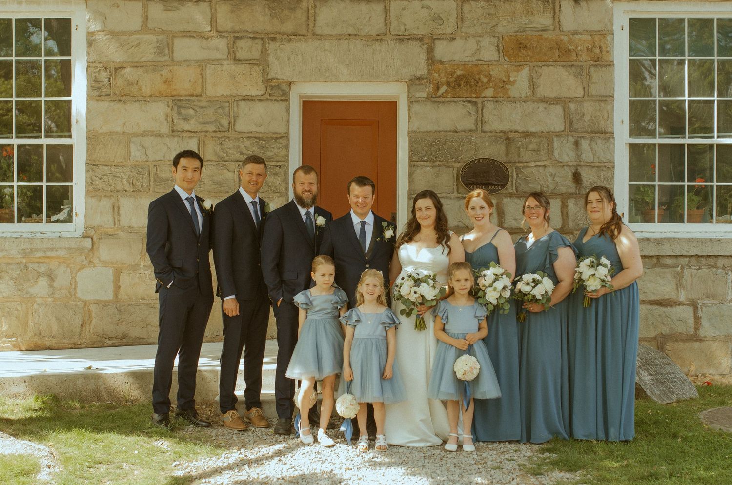 Wedding party poses formally outside stone building with bridesmaids in blue dresses.