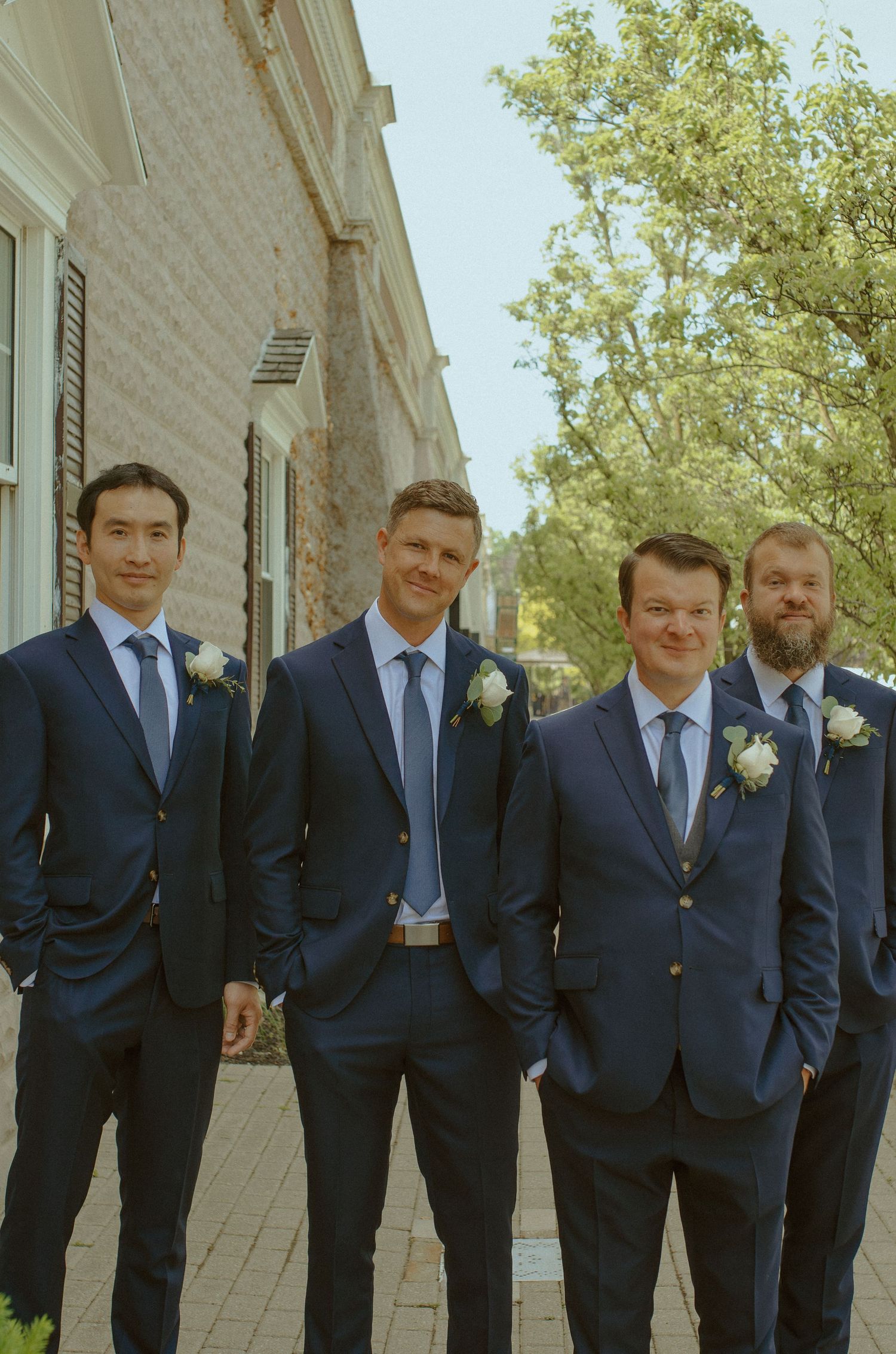Groomsmen in matching navy suits stand in line against light colored building exterior.