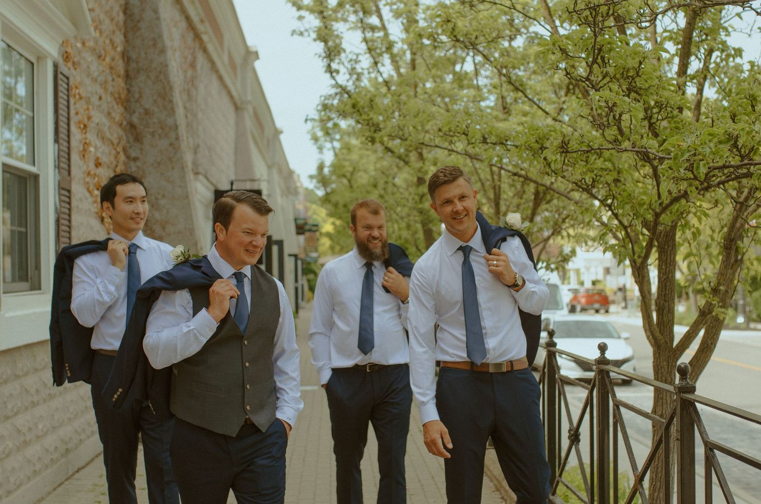 Groomsmen in formal attire laugh and socialize outdoors on wedding day.