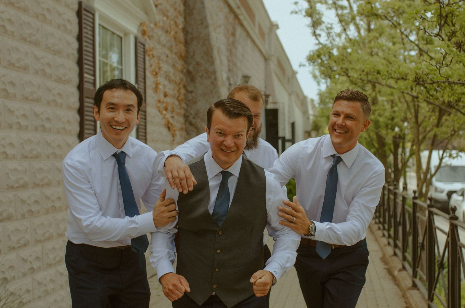Three groomsmen share a playful moment together on a city sidewalk.