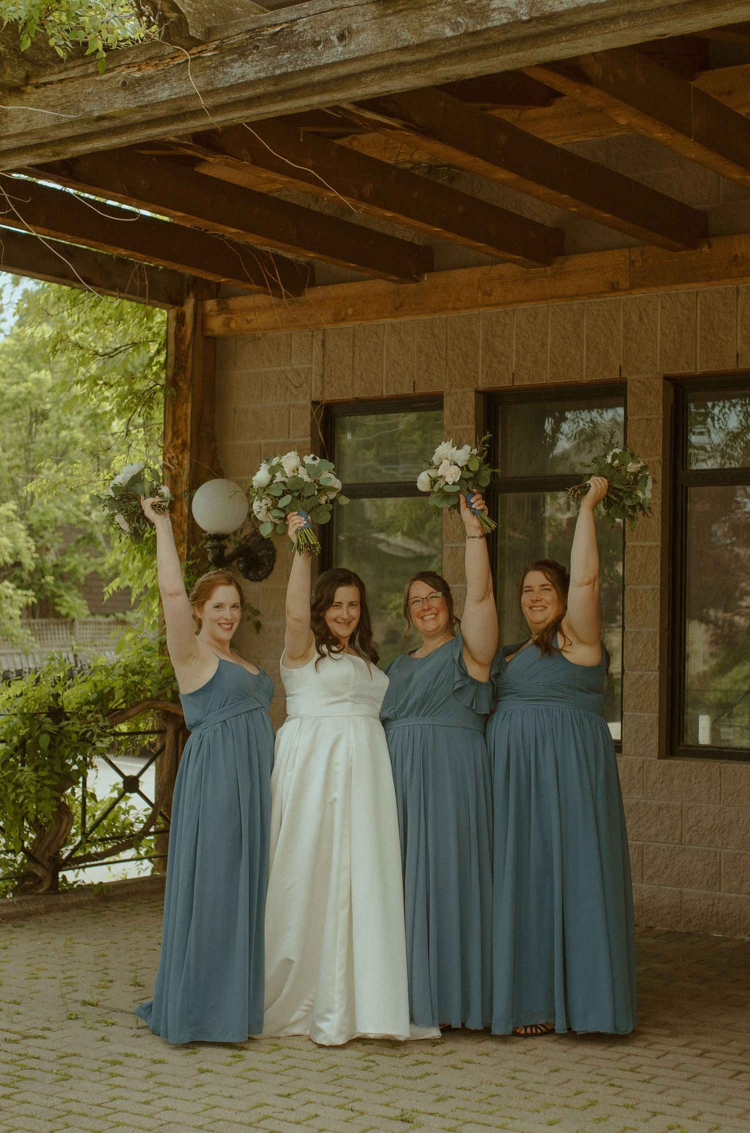 Bridesmaids in dusty blue dresses stand with a wedding gown hung between them under a rustic overhang.