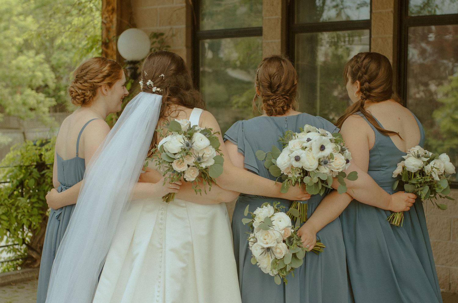 Wedding party members holding white bouquets stand together in light blue dresses.