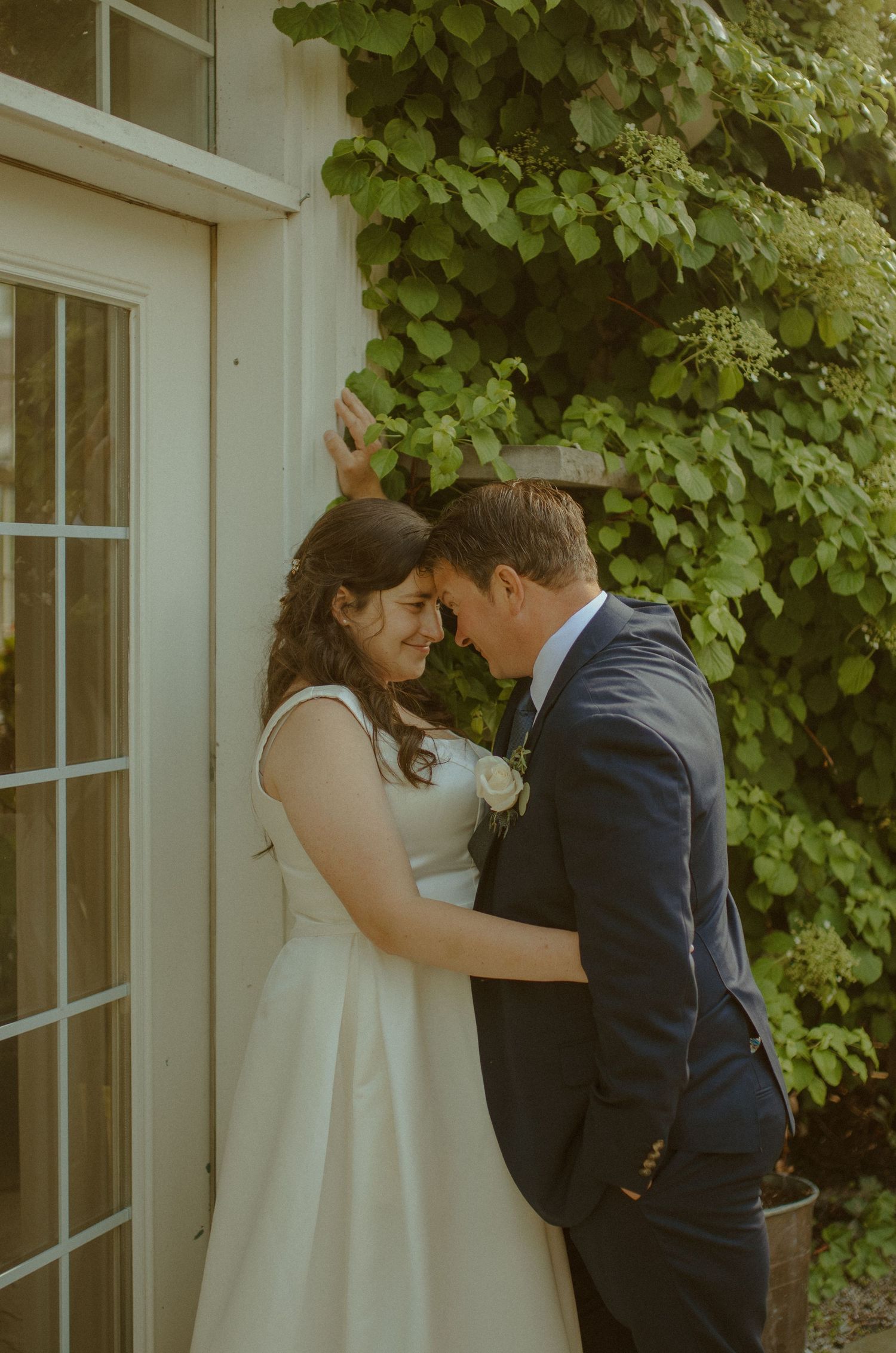A romantic wedding couple shares an intimate moment against a white wall with green ivy.