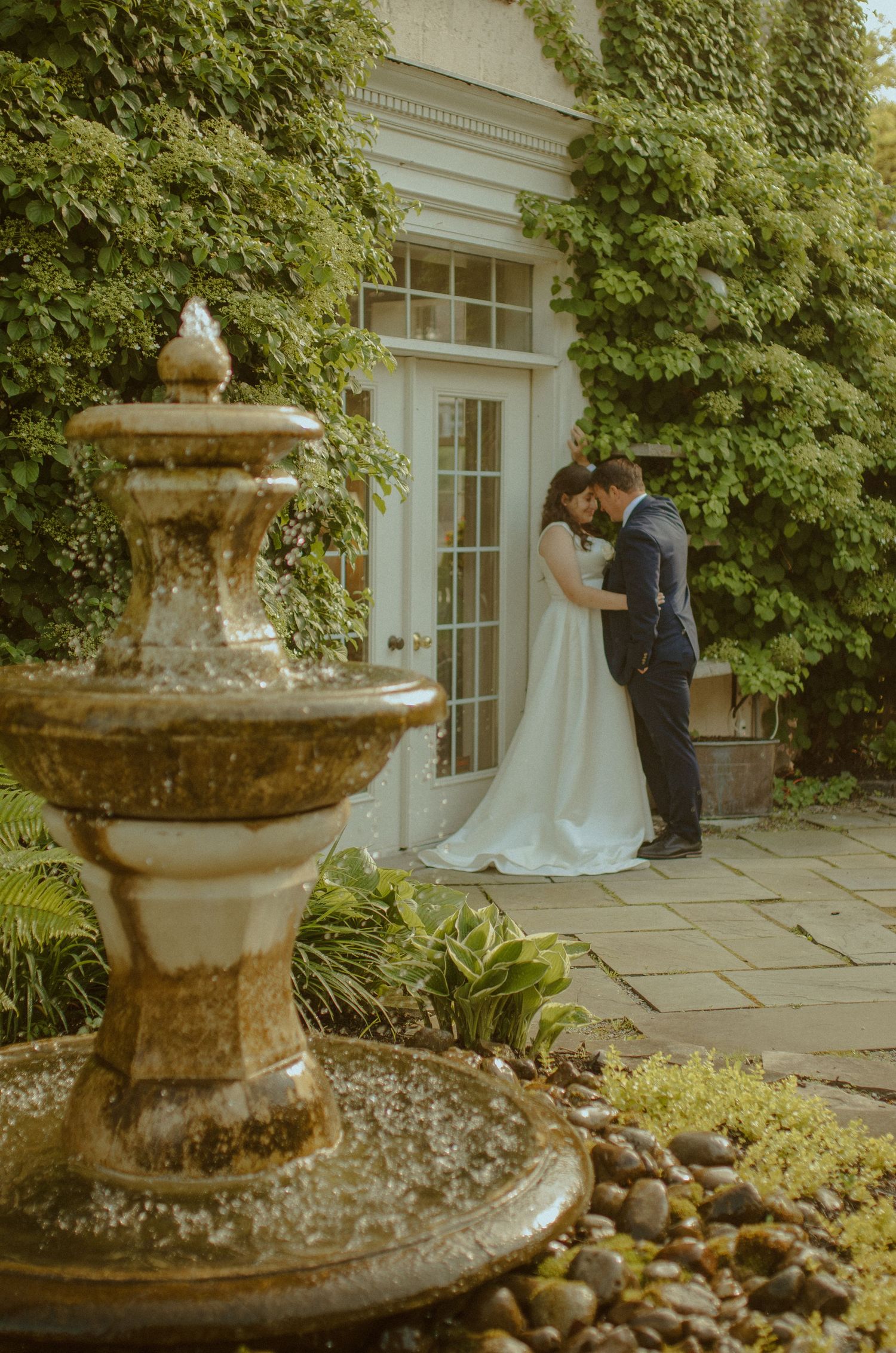 Wedding couple posed by an elegant stone fountain at a formal garden venue.