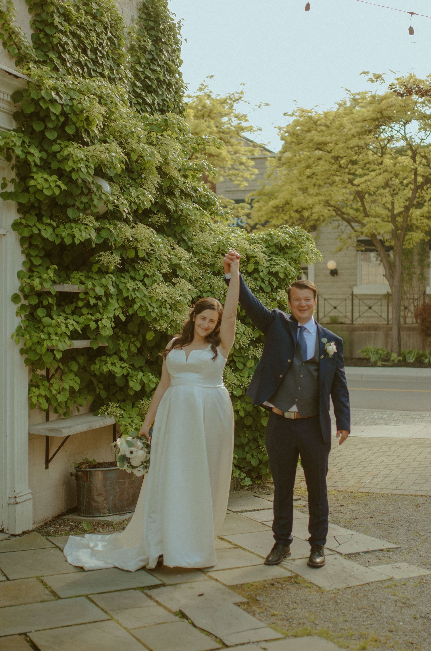 A bride in white and groom in navy raise their joined hands in celebration outside.
