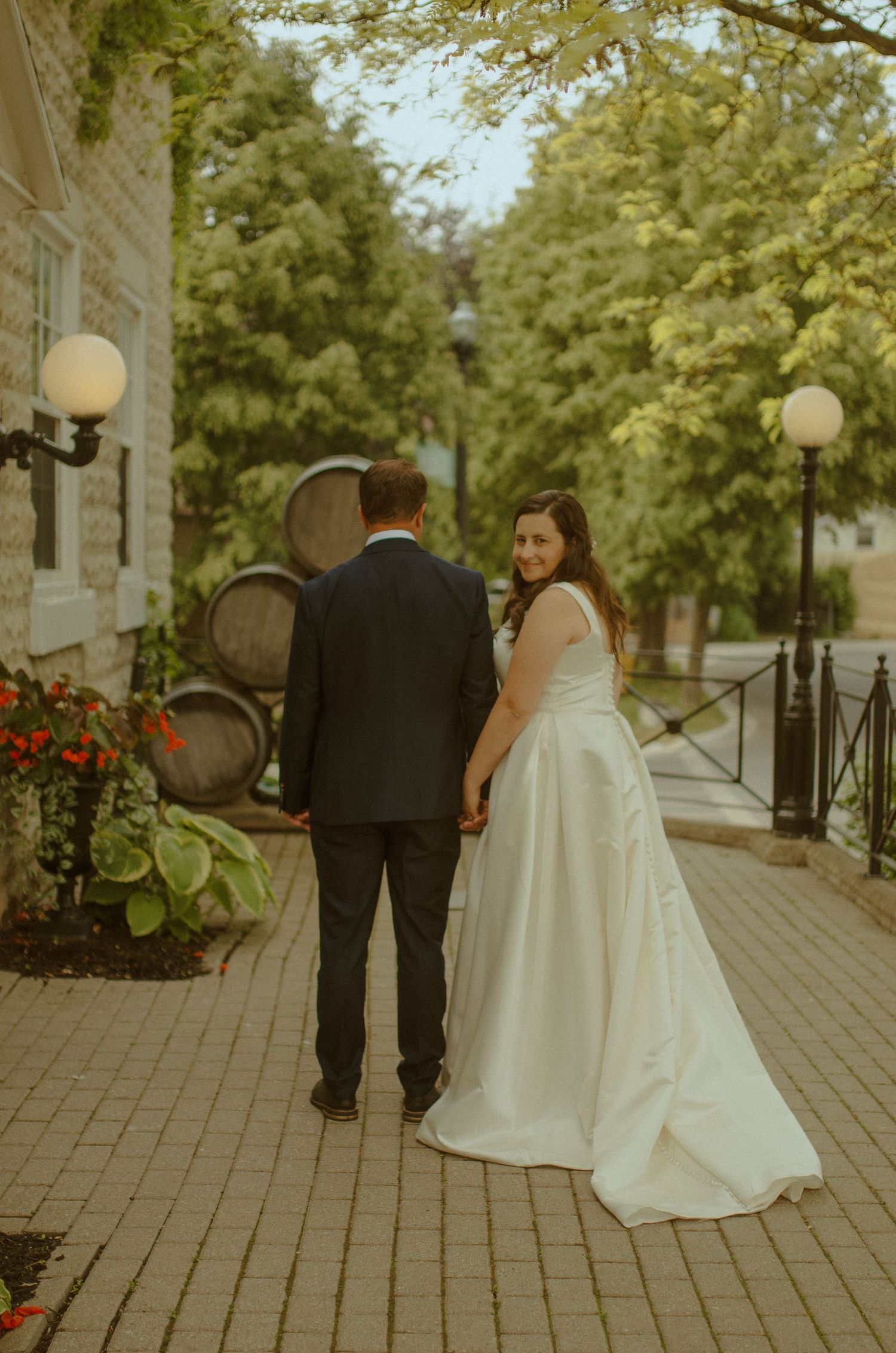 Wedding couple walking together on a brick pathway surrounded by trees and lampposts.