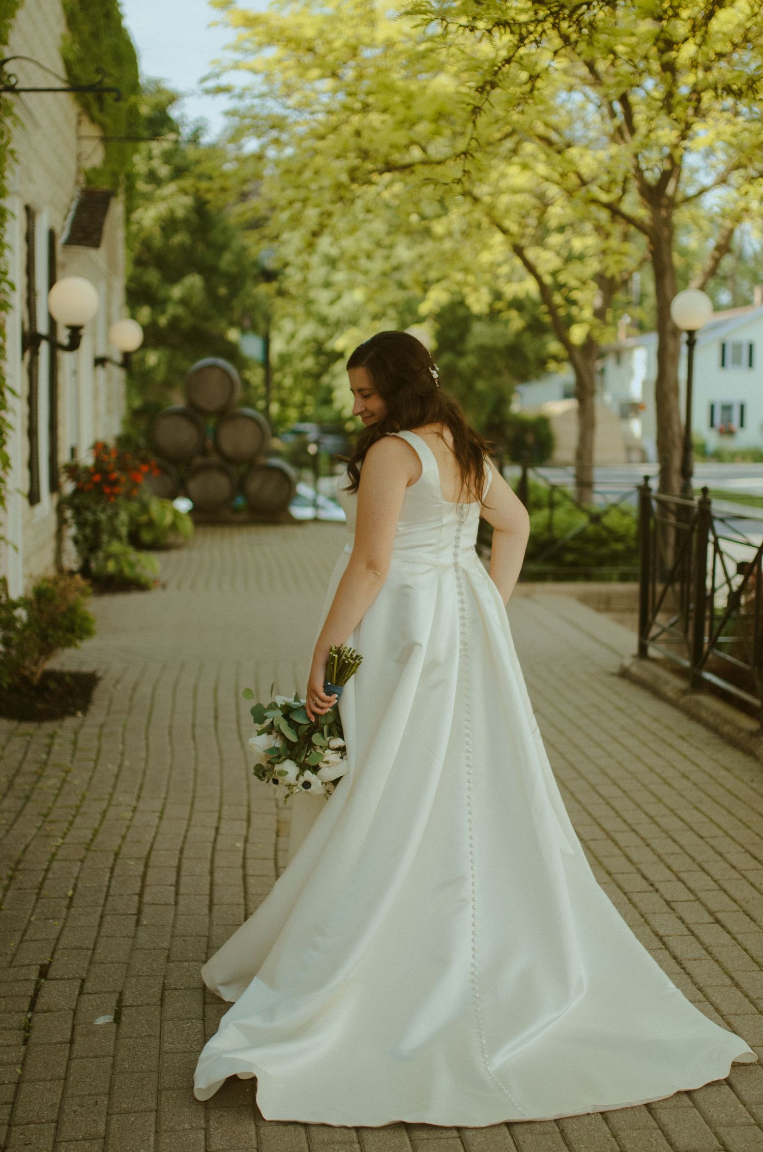 A flowing white wedding dress billows elegantly along a tree-lined brick pathway on a sunny day.