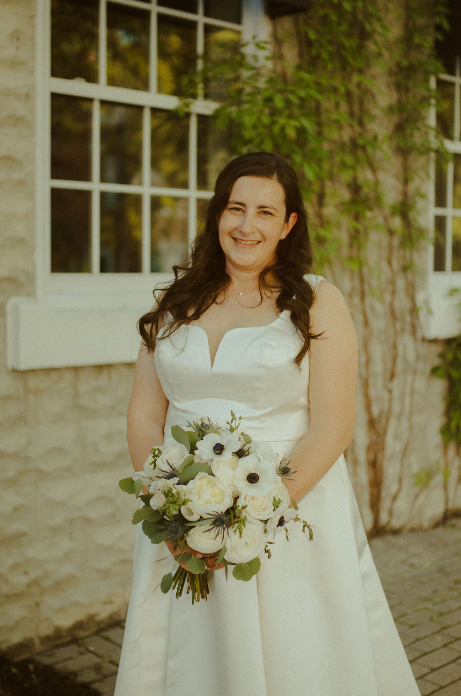 A bride in a white wedding dress holding a delicate bouquet of white flowers and greenery against a stone wall.