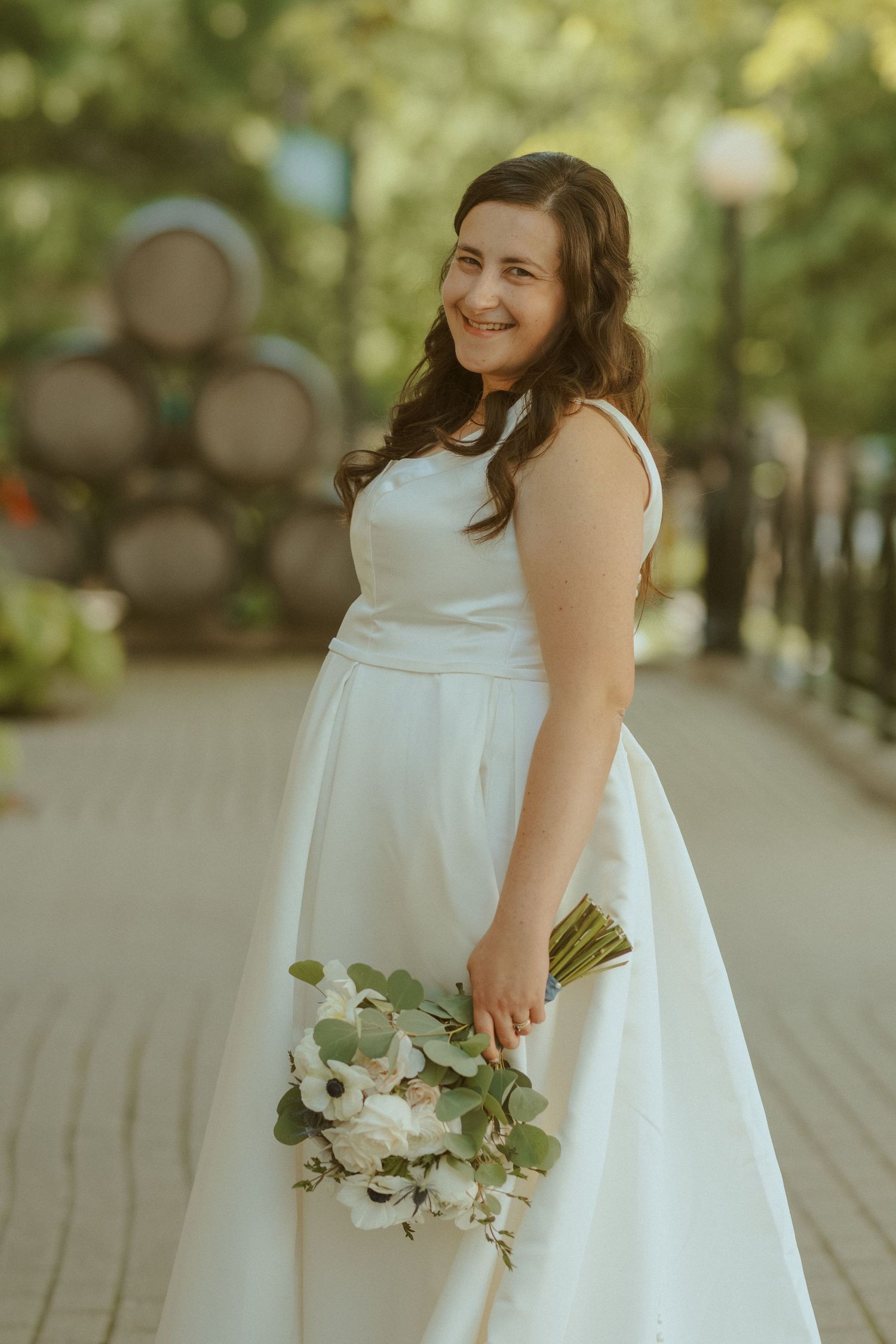A young child wearing a white dress holds a bouquet of greenery while standing on a paved walkway on a sunny day.