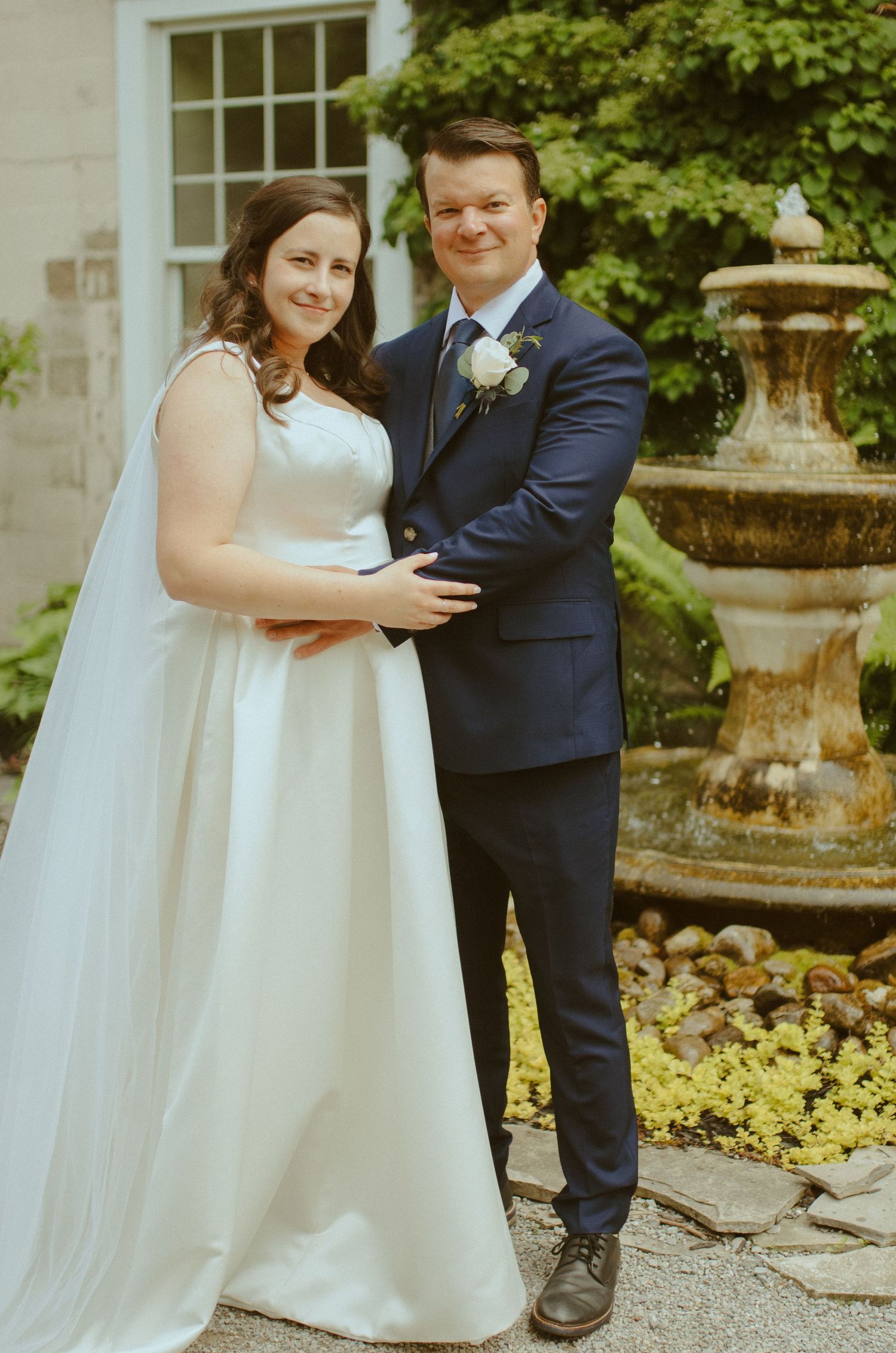 Couple in wedding attire embrace near a decorative garden fountain.