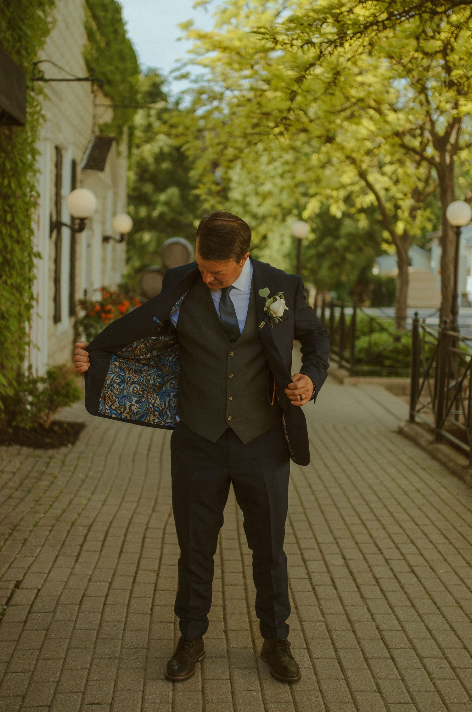 A suited figure shows off the detailed lining of their jacket on a tree-lined brick pathway.