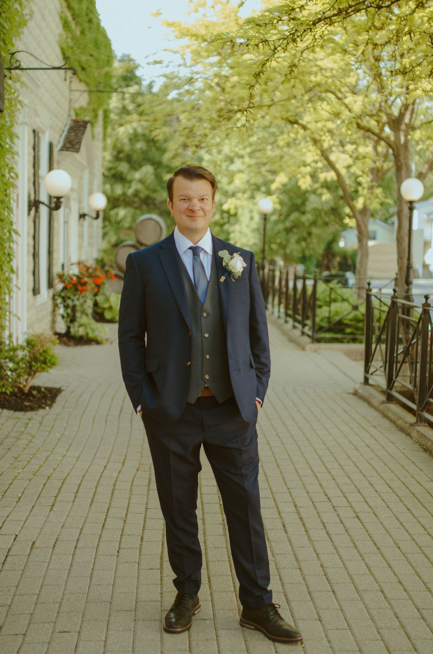 Groom in classic navy suit poses on an outdoor walkway lined with trees and lampposts.