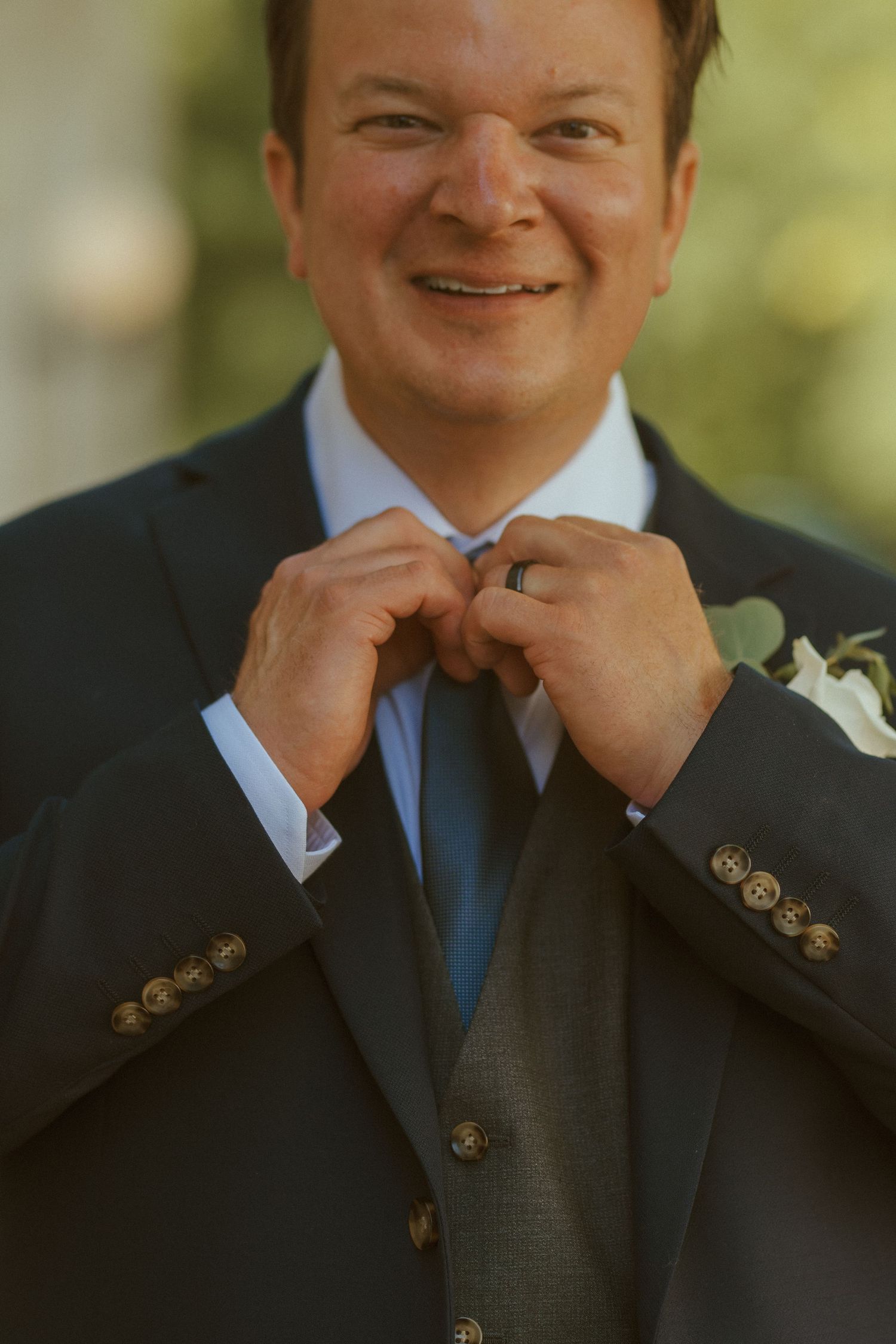 A groom adjusts his tie while wearing a classic black suit and blue tie on his wedding day.