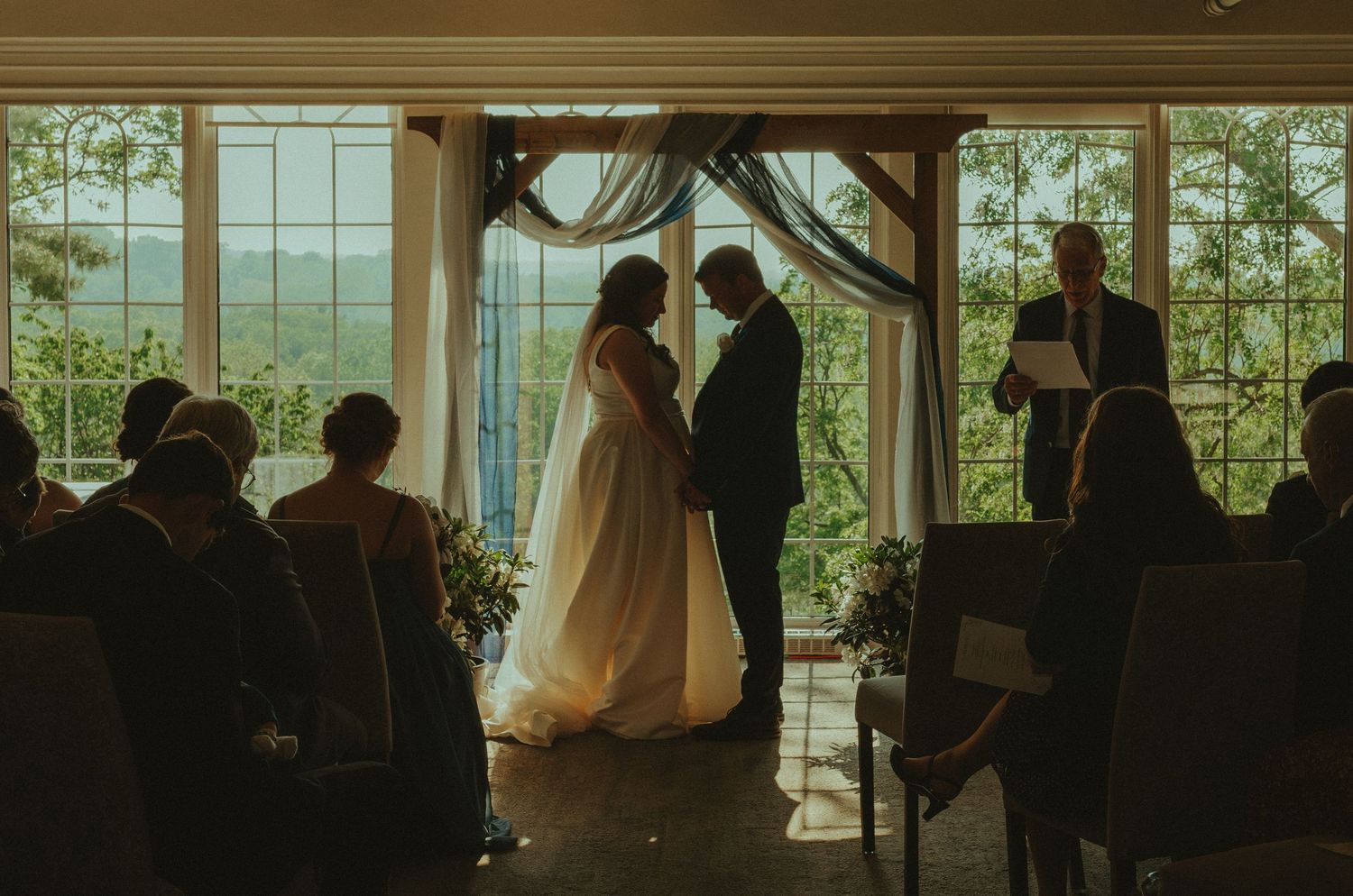 Wedding ceremony silhouette against large windows with mountain views as couple exchanges vows under draped arch.