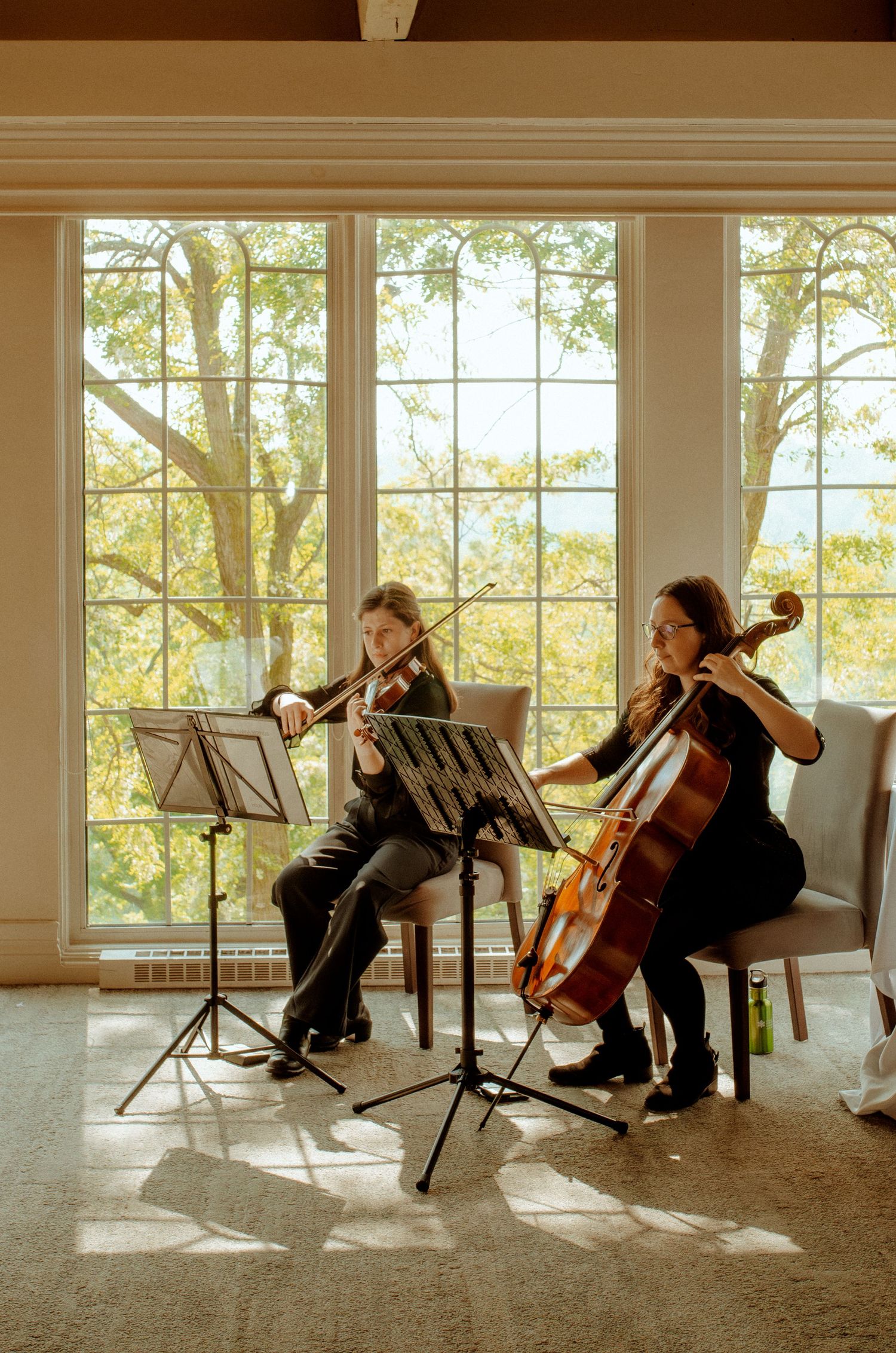 Musicians perform on violin and cello by sunlit windows during wedding ceremony.