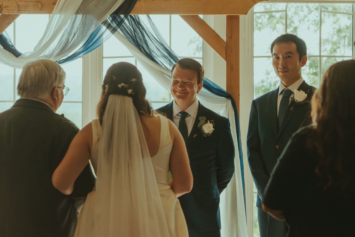 Wedding ceremony inside a bright venue with white drapery and wooden beams.