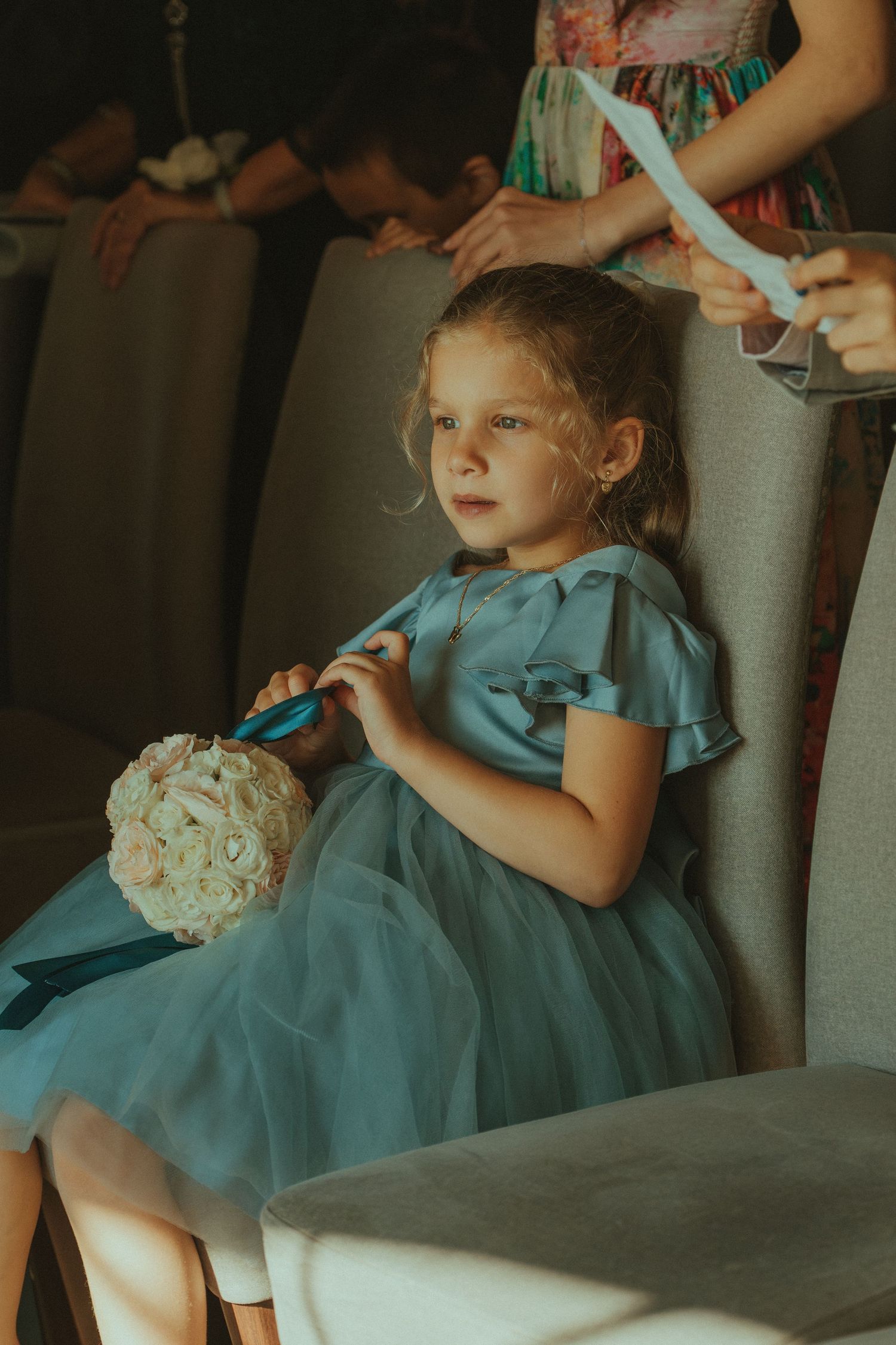 Young attendant in teal dress rests on couch while holding a small white bouquet.