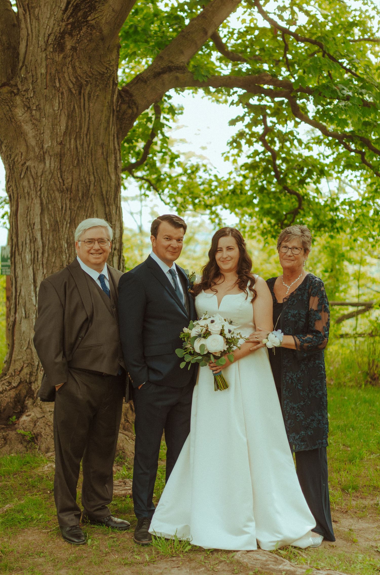 Wedding party poses together under large trees in natural outdoor setting.