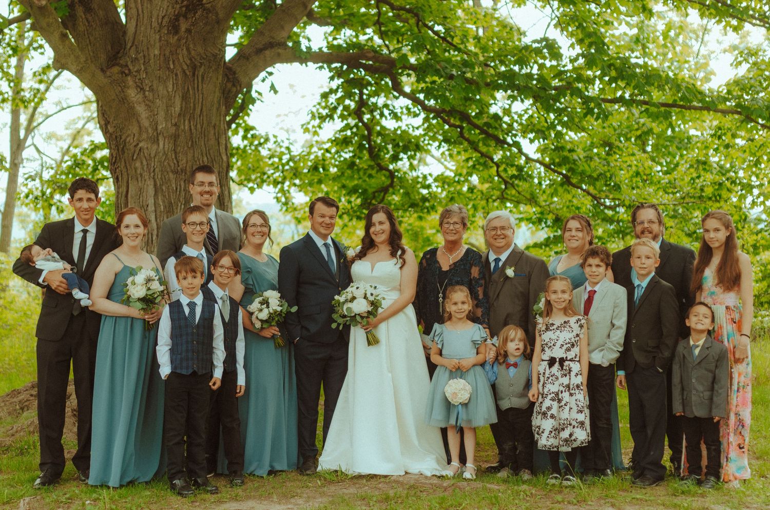 Large wedding party group photo outdoors with bridesmaids in blue dresses under leafy green trees.