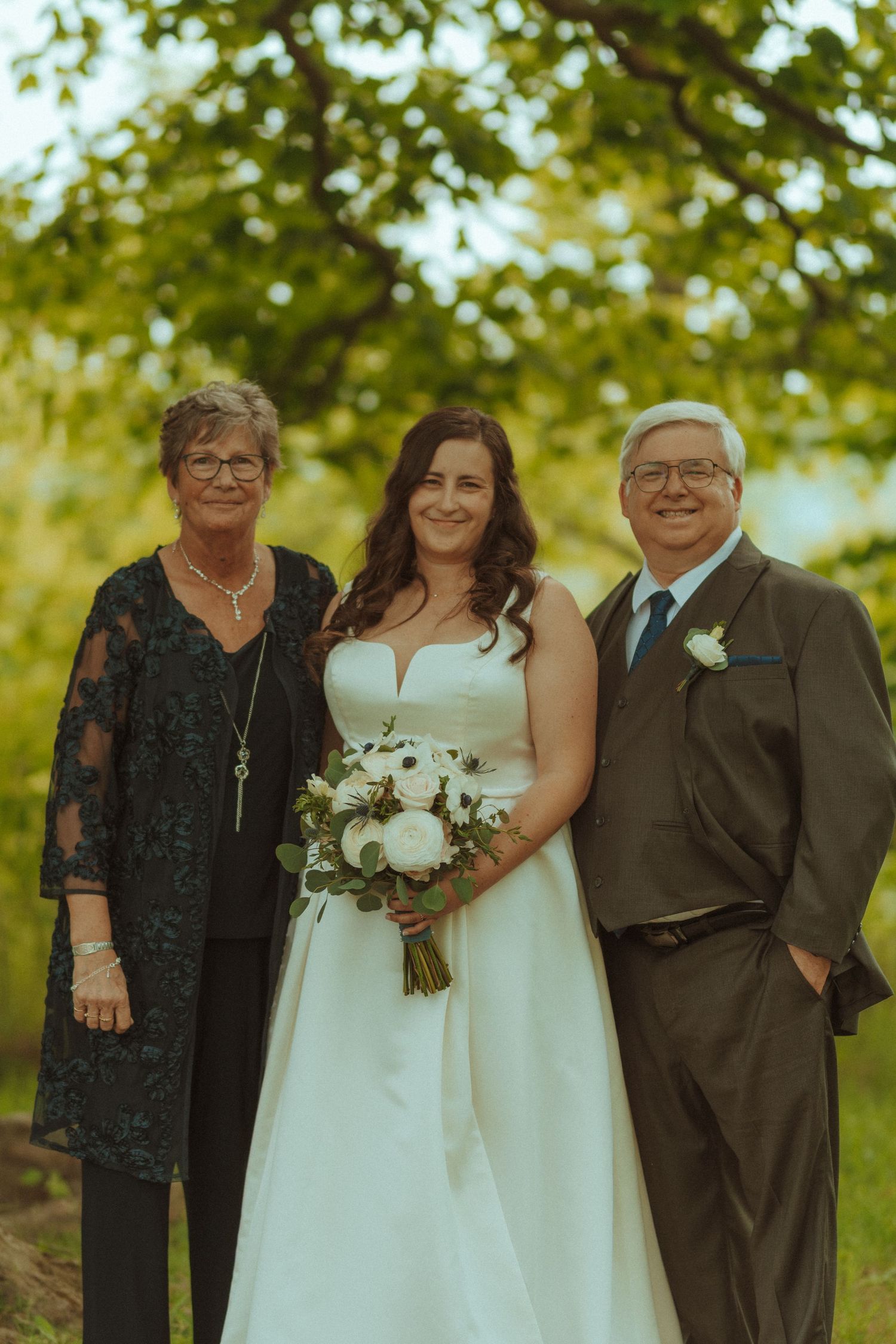 Wedding group photo with family members under leafy green trees.