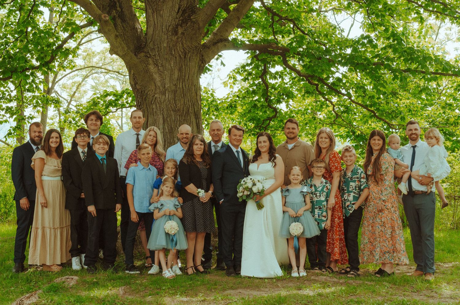 Large group wedding photo of guests gathered under a majestic tree in a garden setting.