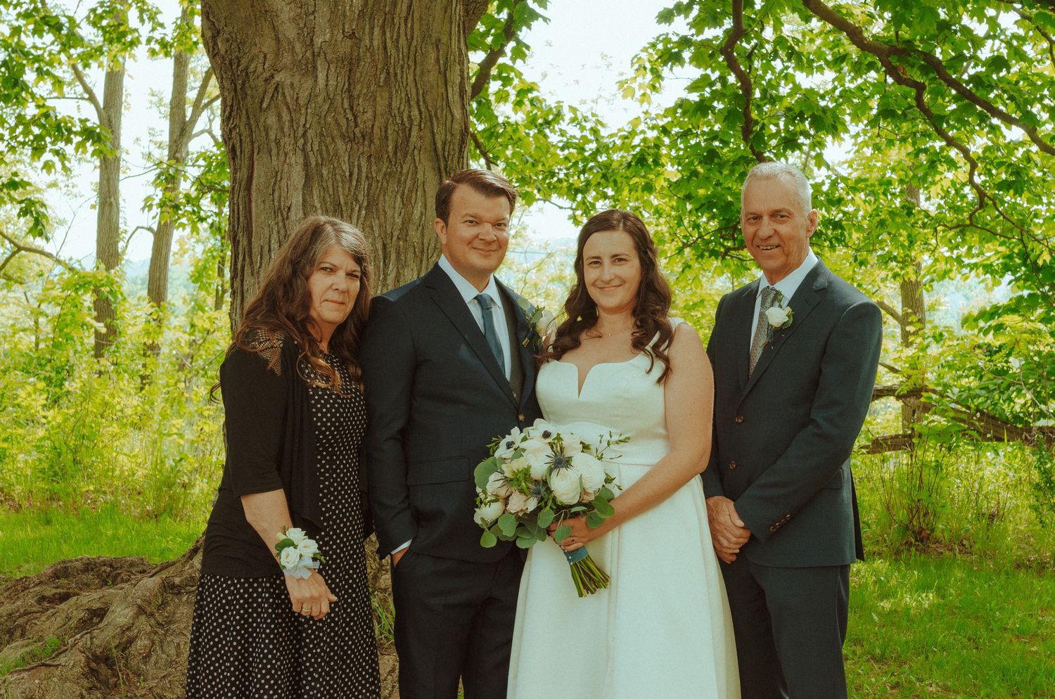 Wedding party poses for group photos under green trees on a bright sunny day with white floral bouquets.