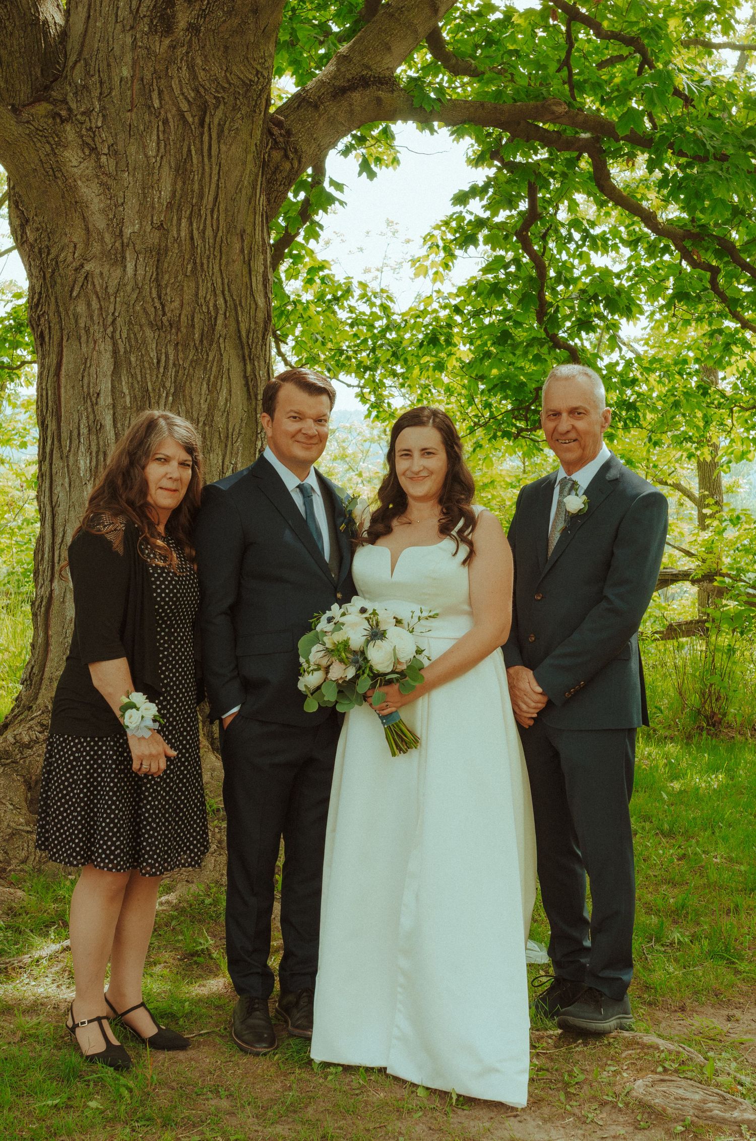 Small wedding group poses for formal portrait under a large tree in natural lighting.
