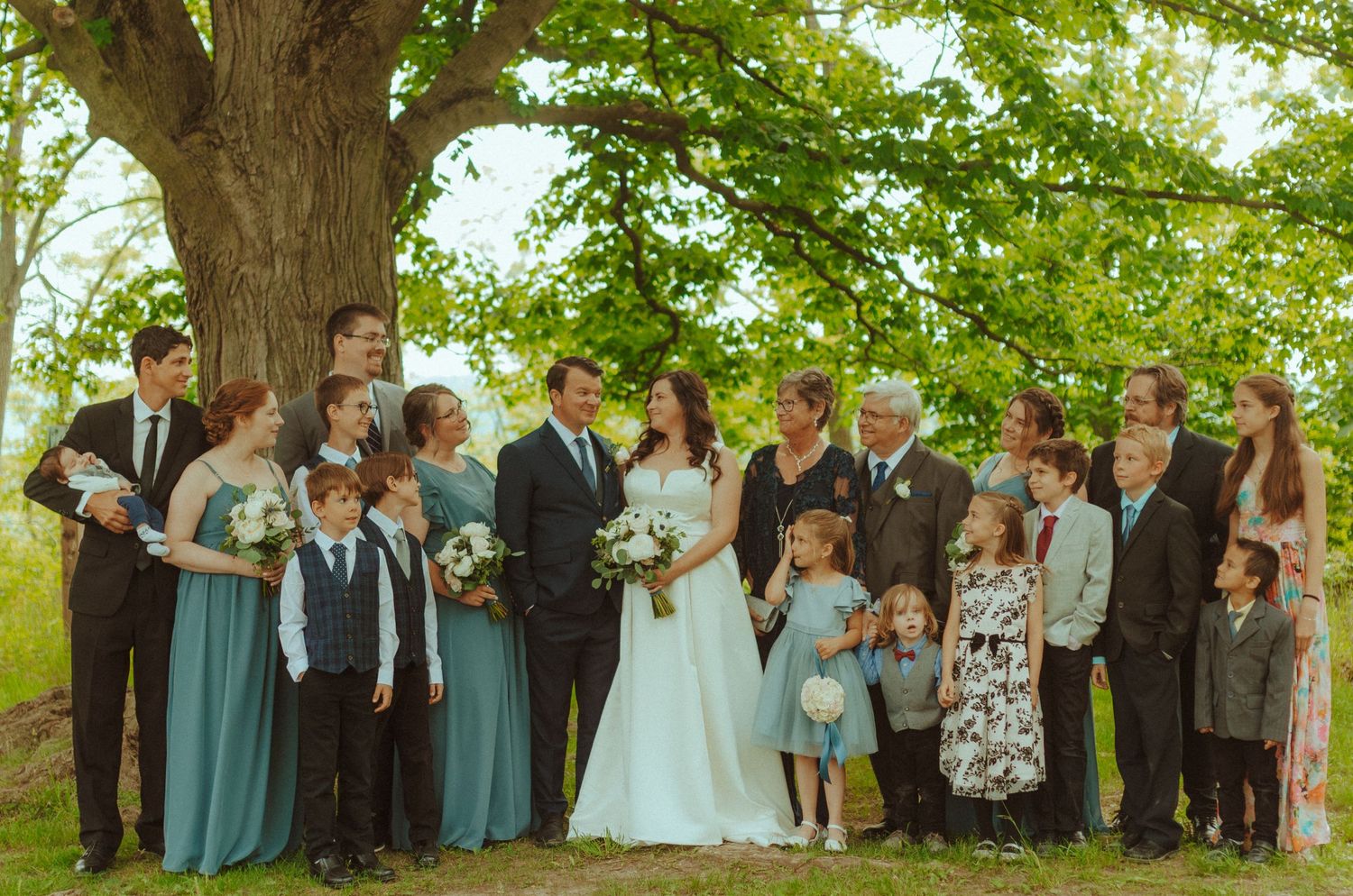 Large wedding party poses together under a big tree for formal group photo.
