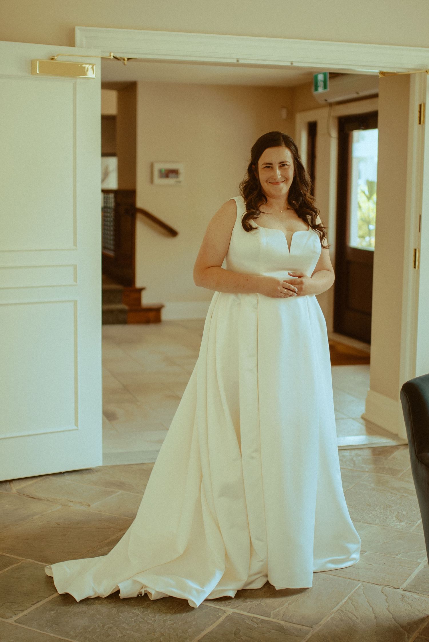A bride in a flowing white wedding dress stands elegantly in a doorway with natural light streaming in.